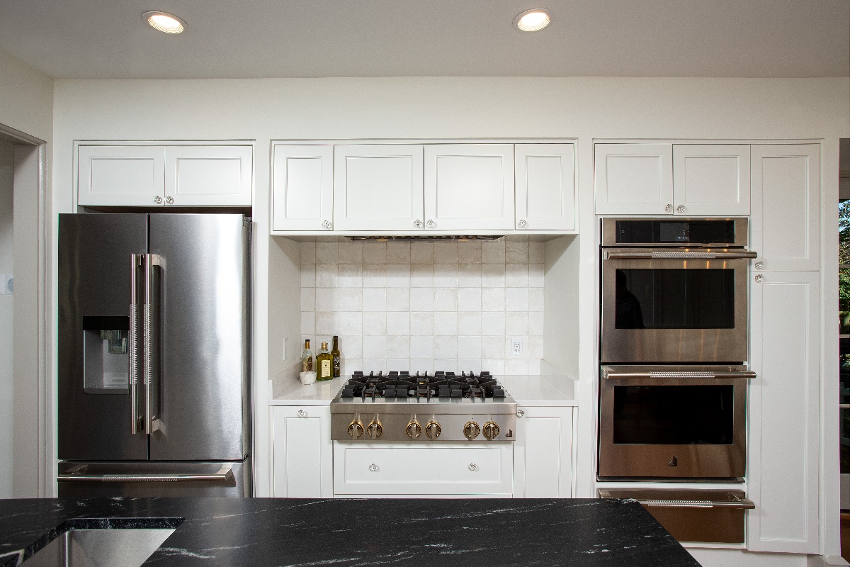 White tile backsplash surrounded by white wood kitchen cabinets, and stainless steel fridge, and ovens built into the wall